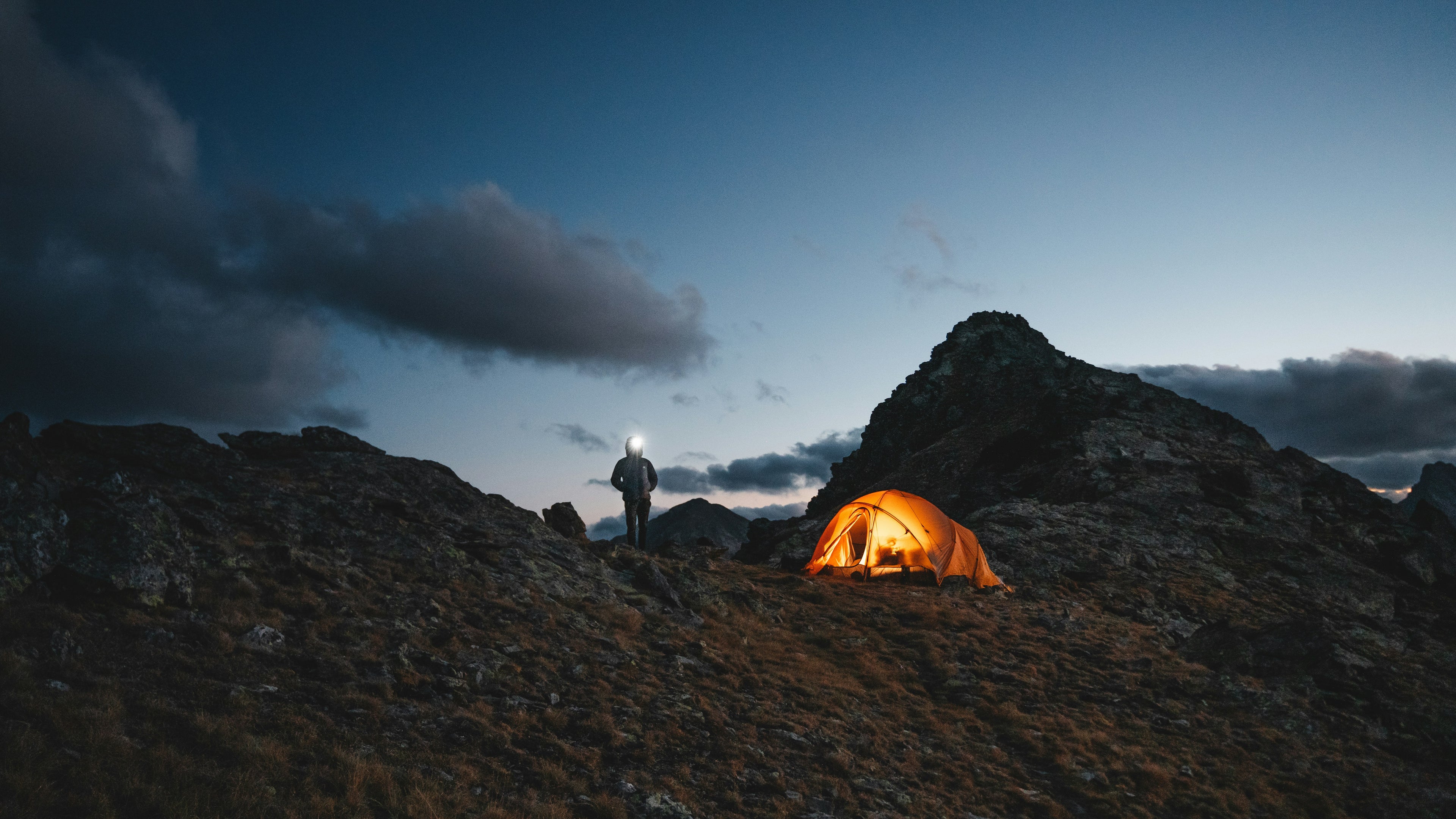 Person with a lantern near a glowing tent on a rocky landscape at night.