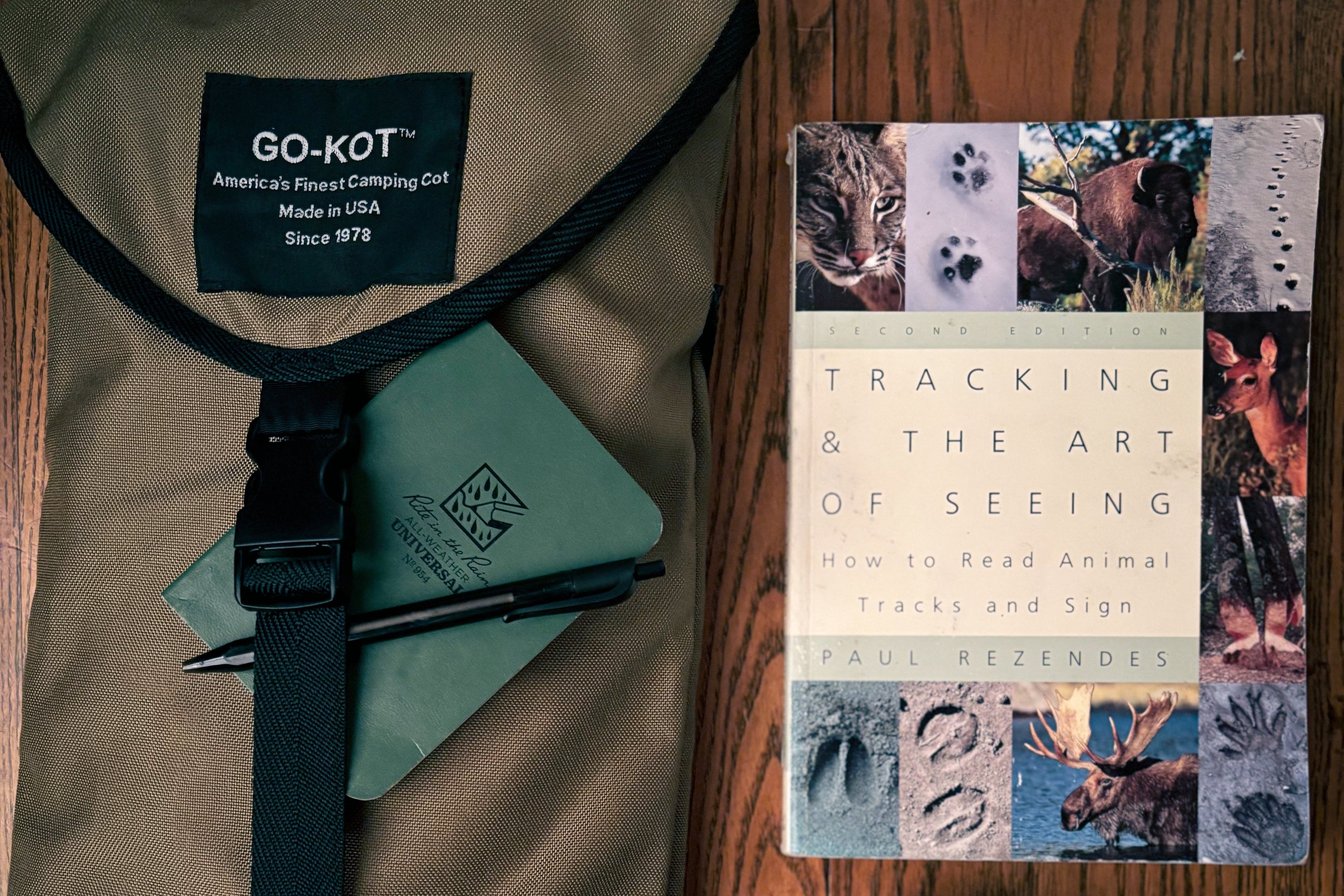 Brown bag with black strap, green notebook, and book titled 'Tracking & the Art of Seeing' on a wooden surface.