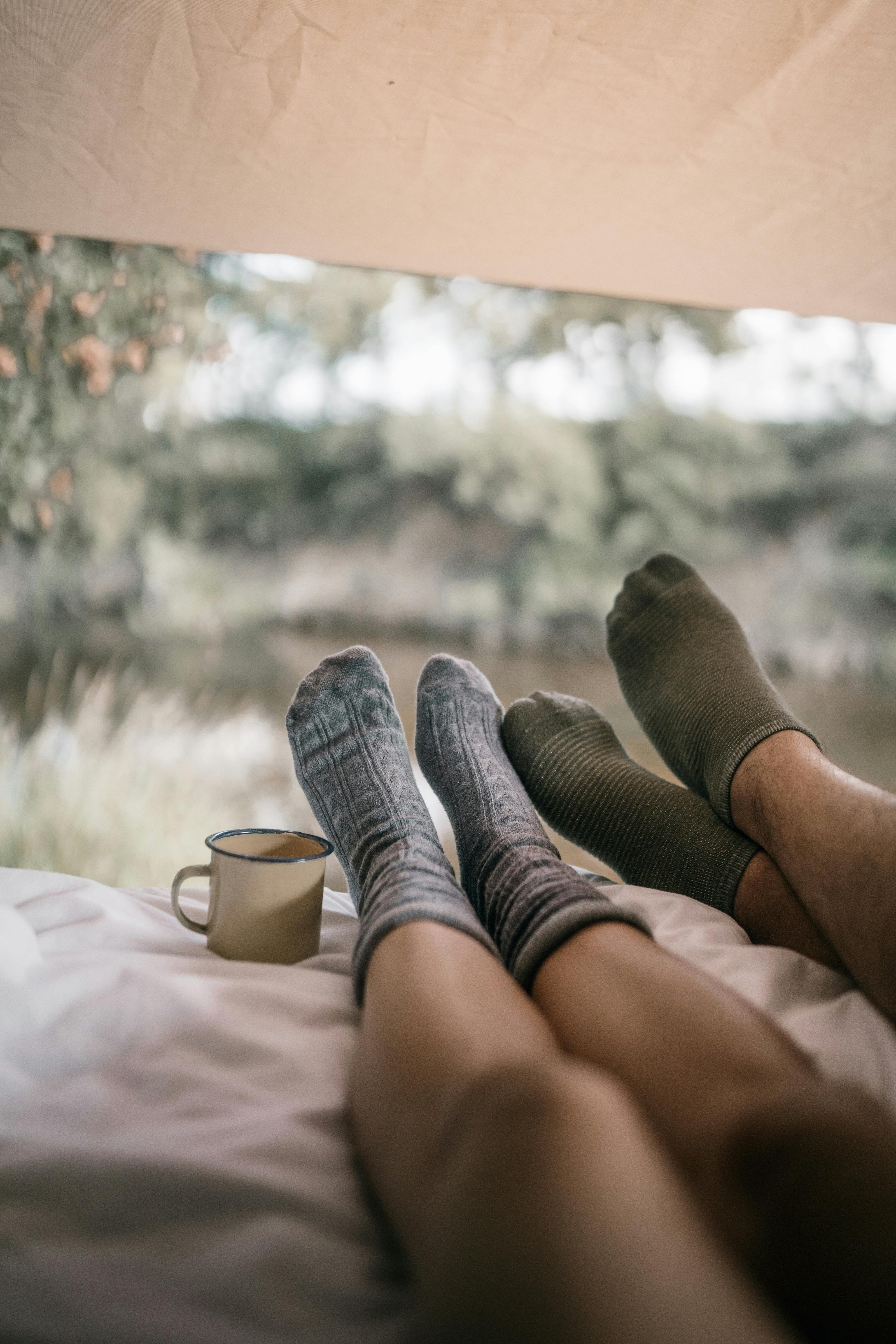 Two pairs of feet in socks with a mug on a bed near a window with a view of trees and water. GO-KOT